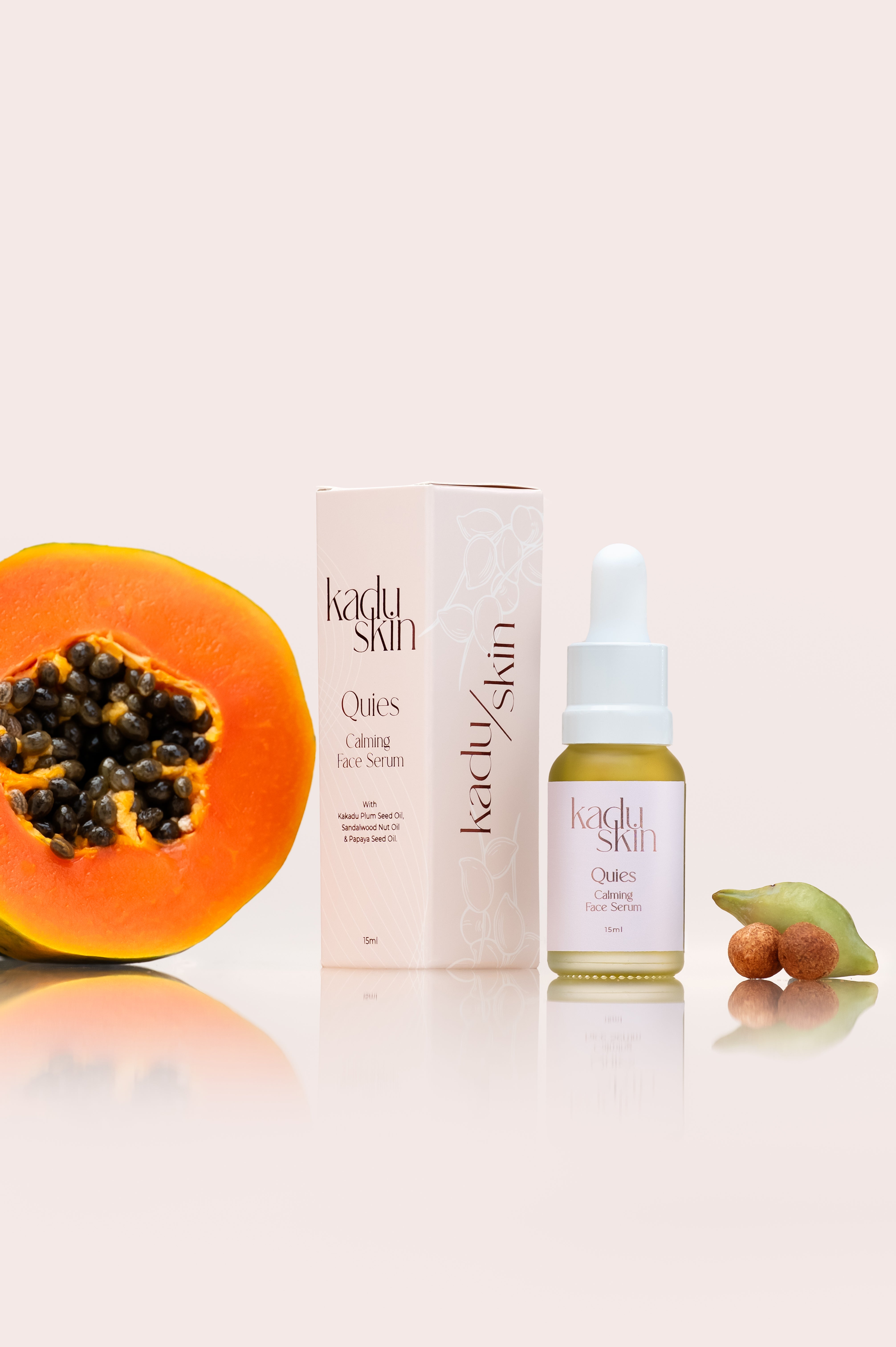 A bottle of Quies Calming Face Serum with a dropper, alongside a box, a halved piece of papaya, and a sandalwood nut. The background is white and the products are on a reflective surface.