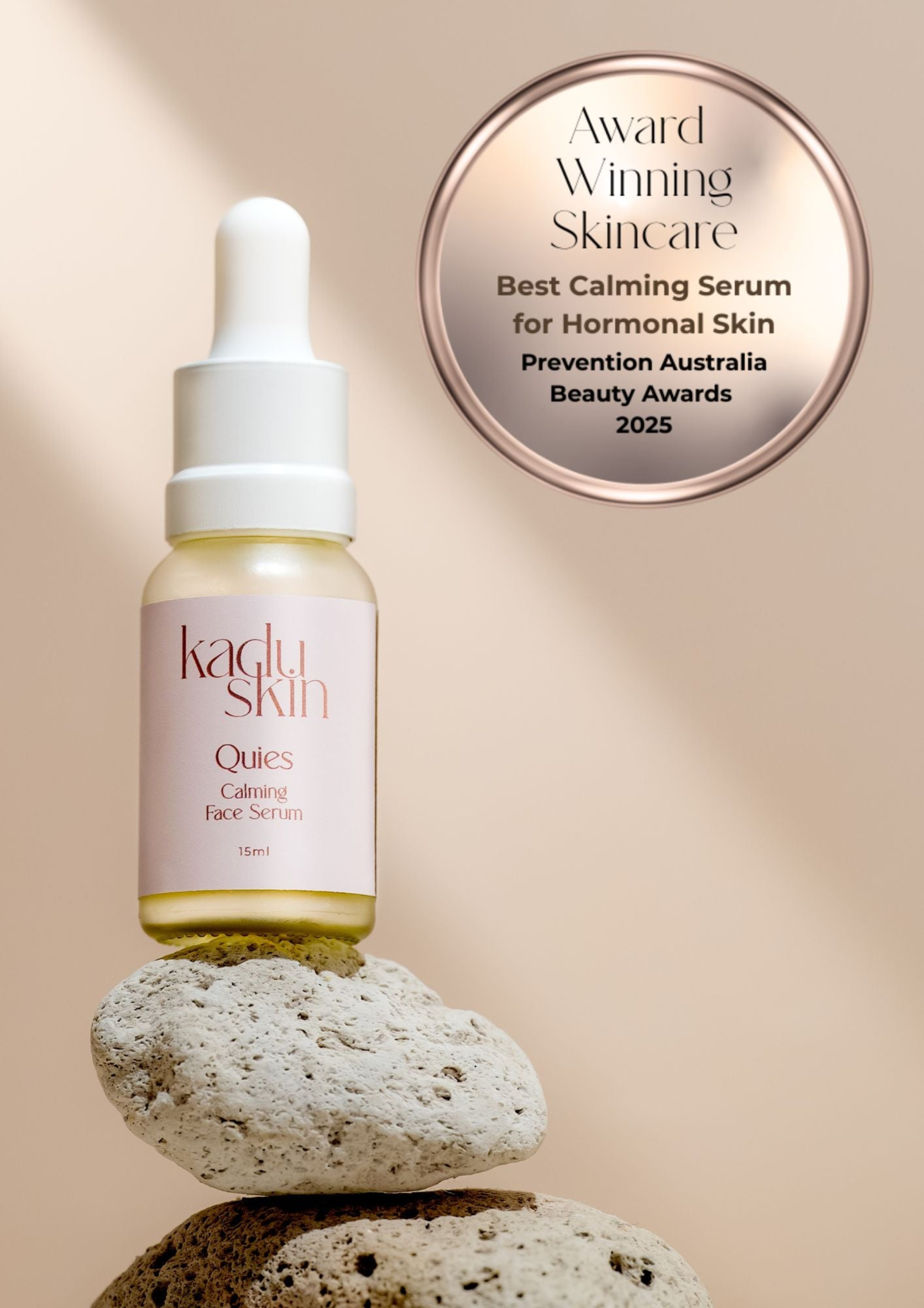 Kadlu Skin serum bottle with dropper on rocks against a beige background, featuring an award badge.